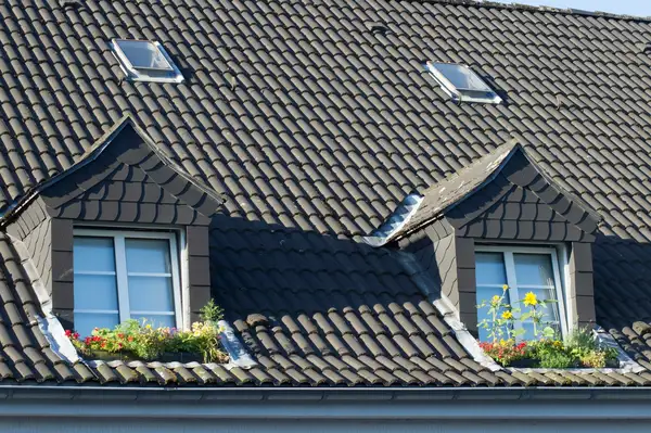 Mörkt tak med takpannor på ett bostadshus med två takkupor och blomlådor under fönstren